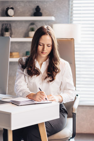 A professional young woman in a white shirt sits at a desk, writing in a notebook. She is focused and engaged, representing productivity, organization, and business planning.の写真素材