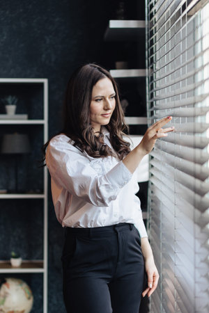 Professional woman in a white shirt stands by the window, adjusting the blinds. She looks outside...