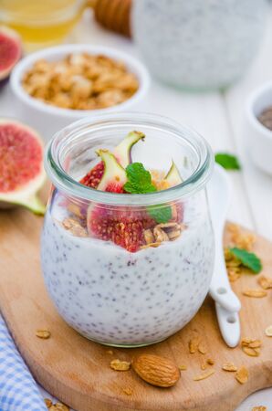Chia pudding with yogurt, homemade granola, fresh figs and honey in a glass jar on a board on a white wooden background.の写真素材