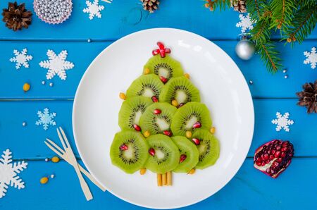 Kiwi Christmas tree on a white plate with pomegranate and sea buckthorn. Christmas food idea. Healthy dessert.の写真素材