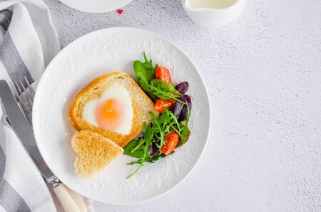 Piece of bread toast cut in shape of heart with egg on a white plate with arugula and cherry tomatoes. Romantic breakfast for Valentine's Day. Top view. Copy space. Horizontal orientation.の写真素材