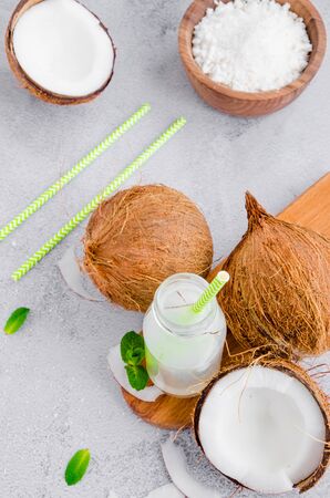 Fresh cold coconut water in a glass bottle with a straw and broken coconuts on a gray background. A healthy drink concept. Vertical orientation. Copy space.