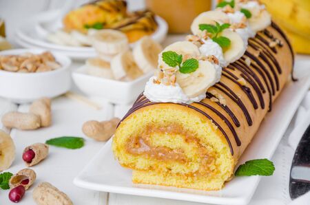 Biscuit or sponge cake roll with peanut butter cream, bananas and chocolate glaze on a white wooden background. Horizontal orientation. Close upの写真素材