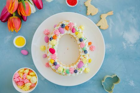 Easter cake egg shaped with cream, candy eggs, cookies, meringue and sprinkles on top on a light blue background with a bouquet of tulips. Easter card. Easter cake. Horizontal, top view, copy spaceの写真素材