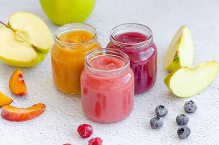 Baby food. Homemade fruit puree. Variety of apple puree or applesauce with frozen peach, raspberries and blueberries in three glass jars on a light background. Healthy food. Horizontal.の写真素材