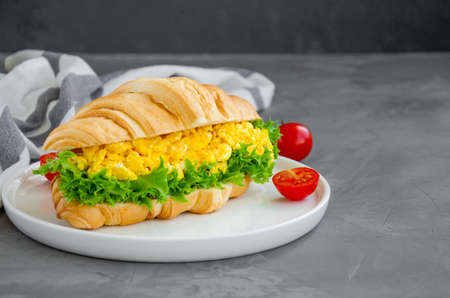 Croissant with scrambled eggs, lettuce and cherry tomatoes on a white plate with a cup of coffee on a dark concrete background. Horizontal, copy spaceの写真素材