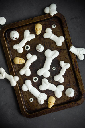 Bones Halloween Meringues with ghosts, chocolate skulls and eyes on an old baking sheet on a dark...