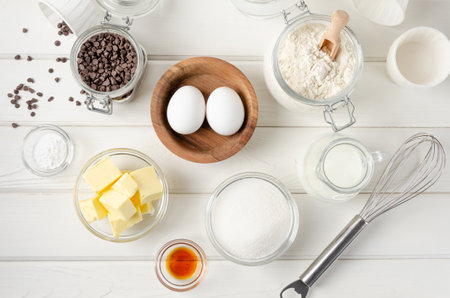 Ingredients for making muffins with chocolate chips. Eggs, butter, sugar, flour, vanilla, chocolate chips on a white wooden background. Recipe step by step. Top viewの写真素材