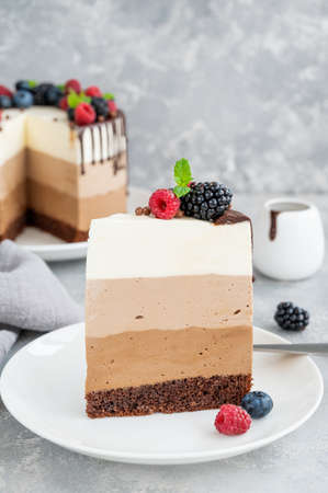 Three chocolate mousse cake with chocolate glaze, blueberries, raspberries and blackberries on top on a white plate on a gray concrete background. Cake with three layers of different taste. Copy spaceの写真素材
