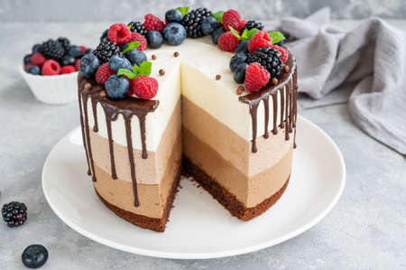 Three chocolate mousse cake with chocolate glaze, blueberries, raspberries and blackberries on top on a white plate on a gray concrete background. Cake with three layers of different taste. Copy spaceの写真素材