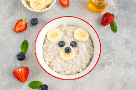 Funny bowl with oat porridge with bear faces made of fruits and berries on a gray concrete background. Food for kids idea, top view, copy space.の写真素材