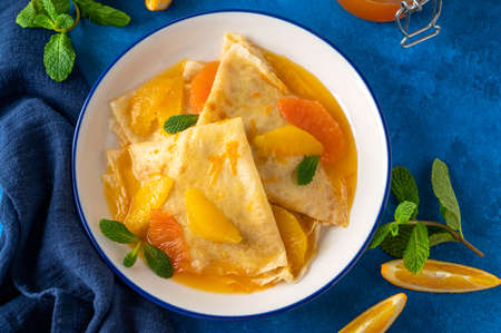 Delicious crepes suzette with orange syrup and slices fruits on a plate on a blue concrete background, top view.の写真素材