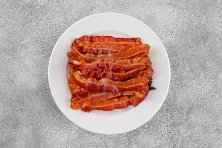 Cooked bacon for breakfast on a white plate on a gray concrete background. Top view, copy space