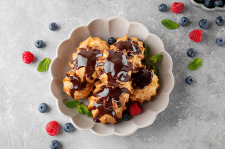 Profiteroles with vanilla cream, fresh berries and chocolate glazed in a bowl on a gray concrete...