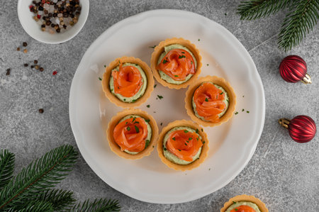 Festive Christmas canapes. Mini tartlets with creamy avocado filling and smoked salmon, garnished with herbs on a white plate on a gray concrete background. Selective focus. Copy spaceの写真素材