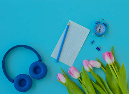 Flat lay home office desk. Workspace with laptop, pink tulips bouquet, headphones, pencil, alarm clock, and a piece of paper for notes on blue background. top view background. spring, international women's day.の写真素材