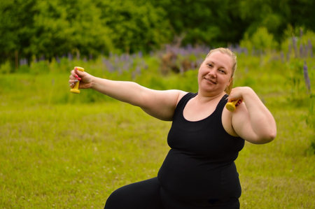 Portrait of cheerful aged plus size woman in fitness wear exercising with yellow dumbbells in park....