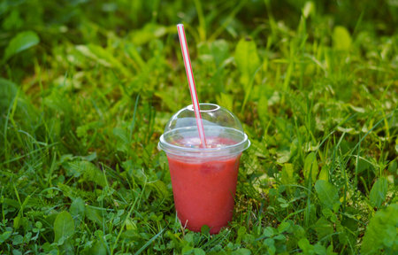 A plastic disposable cup with a pink berry smoothie stands on the grass. Freshness on a hot summer...