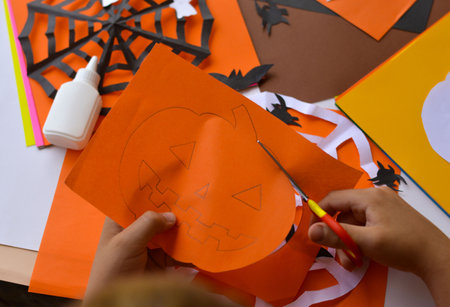 Close-up of children's hands that carves a pumpkin out of orange paper for Halloween. Homemade decorations for the holiday. Hobbies, what to do with children. selective focus.の写真素材