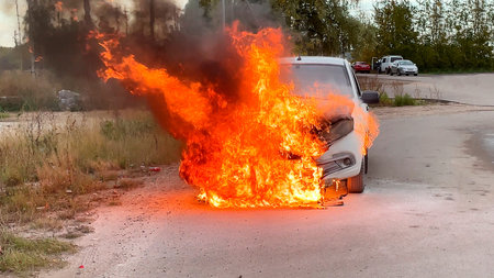 Accident. A white car in an industrial area is on fire. The engine compartment is on fire