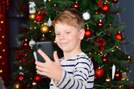 Christmas eve - smiling boy talking photo of themselves on mobile phone. Christmas tree with lights on dark red background.の写真素材