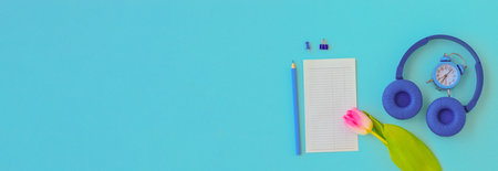 banner flat lay home office desk. Workspace with laptop, pink tulip, headphones, pencil, alarm clock, and a piece of paper for notes on blue background. top view background. spring, international women's day.の写真素材