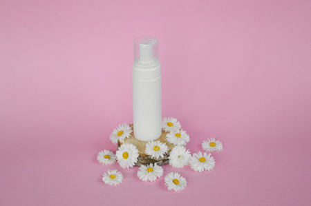 A white plastic jar of cosmetic product stands on a wooden podium with white chamomile flowers on a pink background. Bio cosmetics mock upの写真素材