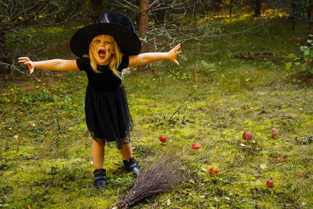 A girl in a witch costume stands with a broom, in an autumn forest. Halloween mysterious lady in woodsの写真素材