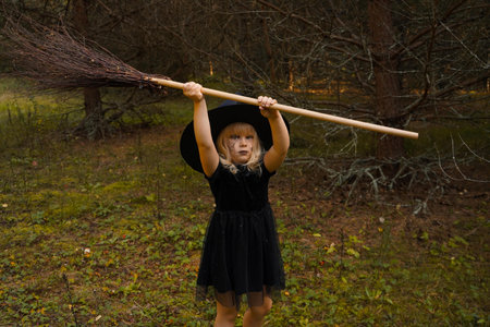 A girl in a witch costume stands with a broom, in an autumn forest. Halloween mysterious lady in woodsの写真素材