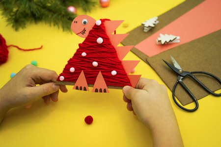 Close-up of hands with Christmas tree toy a dinosaur made by a child out of cardboard and red...