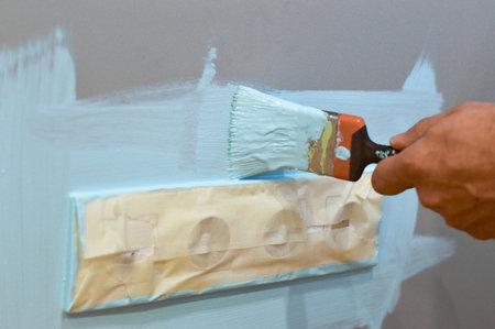 Painting the wall near an electrical outlet with a brush, utilizing masking tape to shield the outlet, during a home renovation task, focusing on neatness and interior maintenance.の写真素材