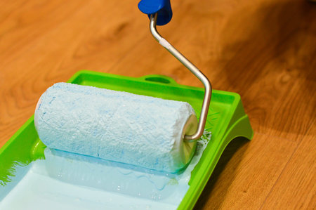 As part of a renovation task, a paint roller is being dipped into a blue paint tray, ensuring an...