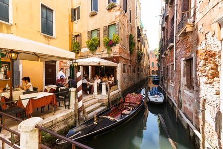 Many visitors in typical tiny cafe in Venice near canal with gondolas and boatsのeditorial素材