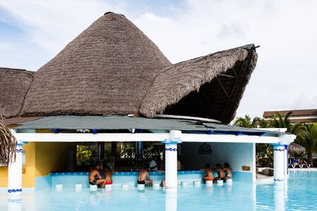 Sun-tanned tourists in swimming suits sitting at the bar in the open pavilion with thatched roof in the middle of the swimming pool.のeditorial素材