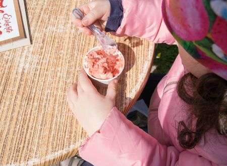 Little child eat pink ice-cream in cap on the table. Two hands of the girl in pink jacket. A ice-cream in spring sun day outside the cafe.の写真素材