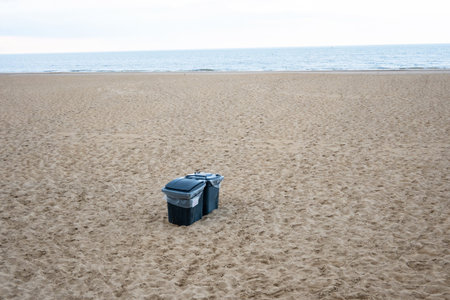 Green and black plastic waste containers buried in yellow sand on the beach. Ostend, Belgium.