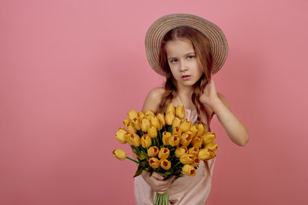 Little girl with pigtails holding flowers wearing hat on pink background. International women's day.の写真素材