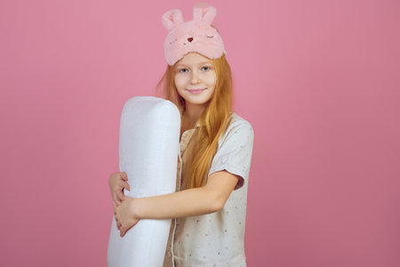 Smiling redhead teen girl holding pillow and wearing sleeping mask on pink background.の写真素材