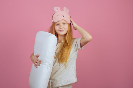 Smiling redhead teen girl holding pillow and wearing sleeping mask on pink background.の写真素材
