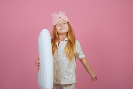 Smiling redhead teen girl holding pillow and wearing sleeping mask on pink background.の写真素材