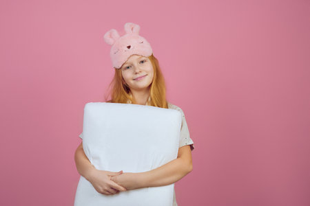 Smiling redhead teen girl holding pillow and wearing sleeping mask on pink background.の写真素材