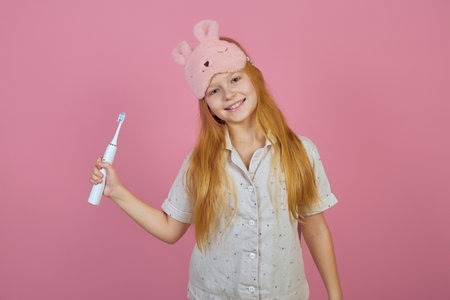 Smiling redhead teen girl holding toothbrush and wearing sleeping mask on pink background.の写真素材