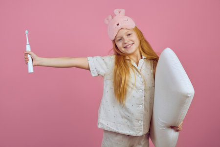 Smiling redhead teen girl holding toothbrush and wearing sleeping mask on pink background.の写真素材