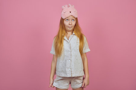 Funny redhead teen girl makes faces wearing sleeping mask on pink background.の写真素材