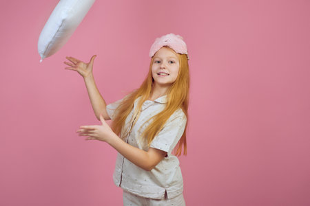 Smiling redhead teen girl throwing pillow and wearing sleeping mask on pink background.の写真素材