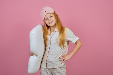 Smiling redhead teen girl holding pillow and wearing sleeping mask on pink background.の写真素材