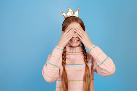 Little smiling girl with golden crown and closed eyes by hands on blue background waiting for surprise.の写真素材
