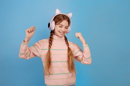 Little smiling girl with headphones on her head and closed eyes dancing on blue background waiting for surprise.の写真素材