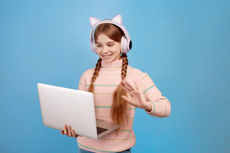 Little girl with headphones on her head have video call with her laptop and waving hand on blue background.の写真素材