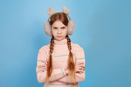 Little angry girl wearing fluffy headphones on blue background. Close portrait.の写真素材